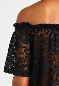 Schwarzes Off-Shoulder-Top aus floralem Spitzenstoff mit einem gewellten Ausschnitt und lockeren, kurzen Ärmeln, das mit filigranen Blumenmustern verziert ist.