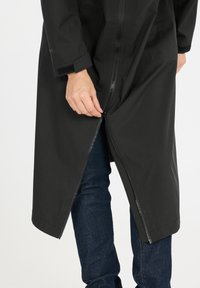 Zwarte waterdichte jas met een ritssluiting aan de voorkant, lange mouwen en een ruime pasvorm. Hand op de ritssluiting, gecombineerd met donkerblauwe jeans.