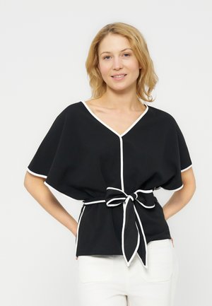 Žena v čiernom topu s véčkovým výstrihom a bielym lemom a opaskom, v bielych nohaviciach, stojaca s rukami za chrbtom.