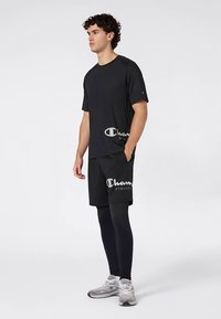 Camiseta deportiva negra con un logo en el dobladillo, combinada con pantalones cortos negros que presentan un logo similar, completada con leggings ajustados negros y zapatillas grises.