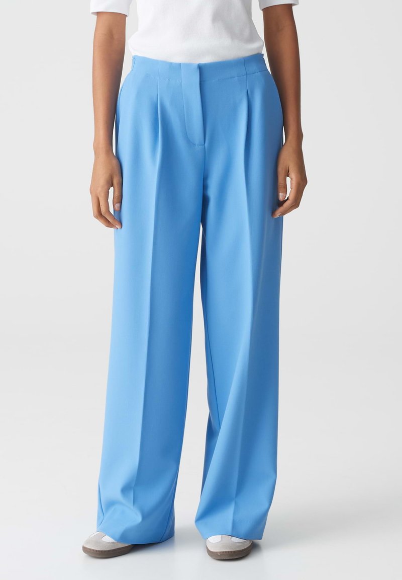 Lichtblauwe wijde broek gemaakt van een gladde stof, met een voornaad en een op maat gemaakte tailleband. Gecombineerd met neutraal gekleurde schoenen.