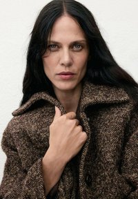 Abrigo de lana tejido en marrón con un amplio cuello, que presenta un patrón texturizado y grandes botones, llevado con una mano sujetando la tela.