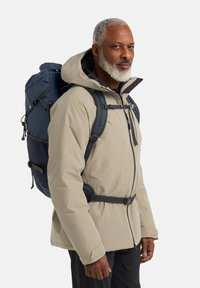 Giacca isolata beige con cappuccio, dotata di cerniere nere e cinturini regolabili, indossata sopra uno zaino blu navy con dettagli in rete.