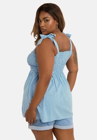 Helles blaues gepunktetes Top mit gerafftem Oberteil, verstellbaren Schulterträgern mit Schleife und einem fließenden Design, kombiniert mit hellen Denim-Shorts.