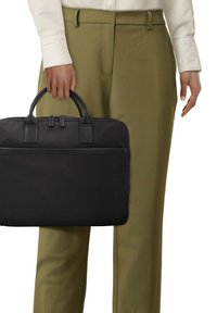 Bolsa de ordenador portátil de nylon negra con asas de cuero texturizado y cremallera, con un diseño delgado y un bolsillo frontal, sostenida al lado de unos pantalones de color verde oliva.