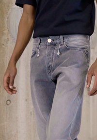 Jeans em denim cinza desbotado com um corte slim, apresentando um fecho de botão e detalhes em corrente decorativa nos bolsos da frente.