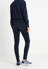 Jeans ajustados de mezclilla azul oscuro con diseño de cinco bolsillos, combinados con zapatillas azul y blancas, y una camisa oscura abotonada, vistos desde atrás.