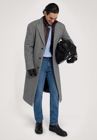 Šedý vlněný kabát s límcem, hnědá kožená bunda pod ním, modrá pruhovaná košile, džíny a černá obuv. Černá taška v ruce.