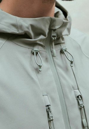 Gros plan sur une personne portant une veste gris clair avec un col montant, une fermeture éclair et des cordons réglables sur la capuche et les poches poitrine.