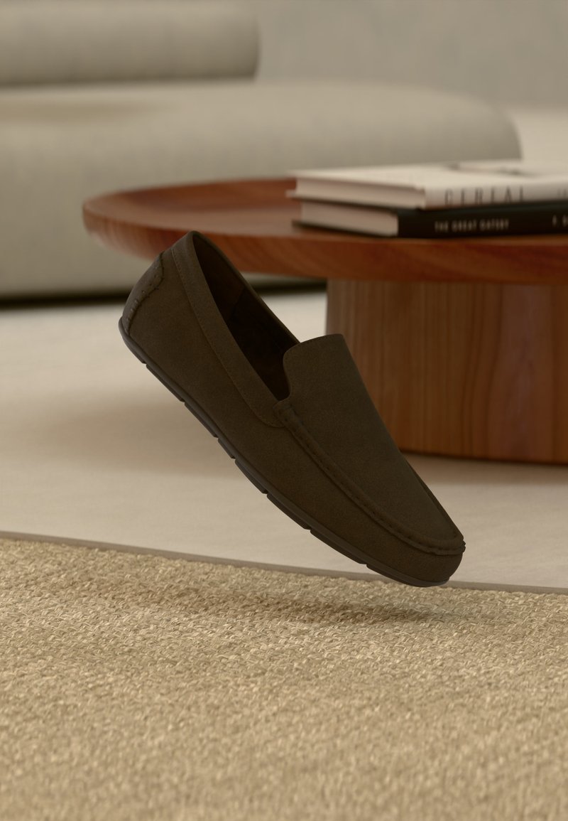 Mocassin marron foncé à enfiler flottant au-dessus d'un tapis beige texturé, avec une table basse ronde en bois et des livres flous en arrière-plan.