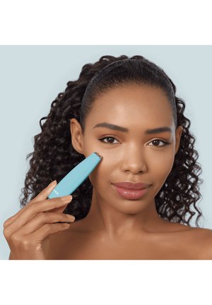 Kvinde med krøllet hår bruger en lille blå ansigtsenhed på sin kind mod en lyseblå baggrund.