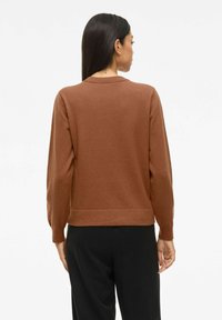 Femme aux longs cheveux bruns portant un pull marron à manches longues et un pantalon noir, vue de dos sur un fond uni.