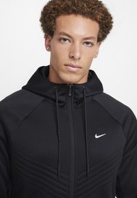 Zwarte hoodie met een textuurontwerp, voorzien van een ritssluiting, aansnoerkoorden en een klein wit Nike-logo op de borst.