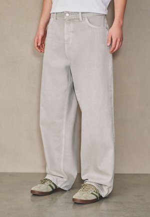 Persoon draagt losse lichtgrijze broek en beige sneakers met groene strepen, staand tegen een effen beige achtergrond.