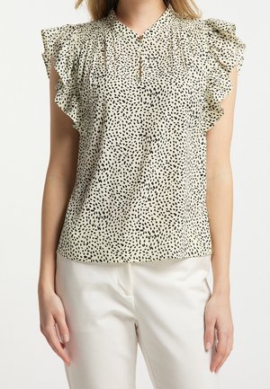 Vrouw draagt een crèmekleurige blouse met een onregelmatig zwart stippenpatroon en gerimpelde mouwtjes, gecombineerd met witte broek.