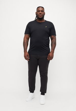 Fekete, rövid ujjú póló arany logóval a mellkasán, fekete slim-fit nadrággal és fehér sportcipővel párosítva. Egyszerű, laza stílus.
