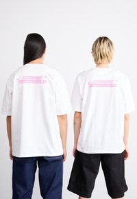 Balti per dideli t-shirt su rožinėmis "adidas originals" užrašu ant nugaros, demonstruojami dviejų asmenų, vilkinčių tamsias šortus.