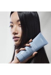 Lichtblauwe tube Aveda Smooth Infusion conditioner vastgehouden tegen een glad, donkerharig gezicht, met een minimalistische achtergrond.