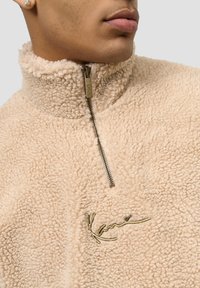 Beige fleecetrøye med høy krage og halv glidelås. Har brodert logo i gull. Mykt, teksturert materiale med en koselig utseende.