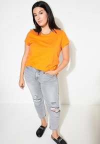 Orange t-shirt med kort ärm och svarta detaljer, matchad med ljusgrå slitna jeans och svarta slip-on-skor med kedjedetalj.