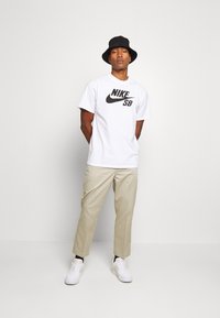 Vit bomull t-shirt med en stor svart Nike SB-logotyp, beige byxor och vita sneakers. En svart bucket hat fulländar outfiten.