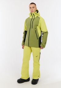 Helle gelbe und grüne wetterfeste Jacke mit einer Reißverschlusstasche; kombiniert mit passenden leuchtend gelben Hosen. Geeignet für Aktivitäten im Freien.