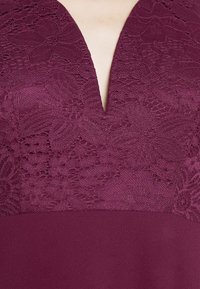 Gros plan d'une robe violette à décolleté en V profond avec des détails en dentelle florale sur le corsage supérieur et un tissu lisse sous la taille.