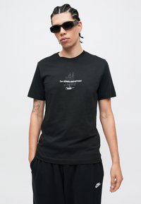 Μαύρο βαμβακερό T-shirt με στρογγυλή λαιμόκοψη, με λευκό κείμενο �και γραφικό σχέδιο. Το μοντέλο φοράει μαύρα γυαλιά ηλίου και χαλαρά μαύρα παντελόνια.