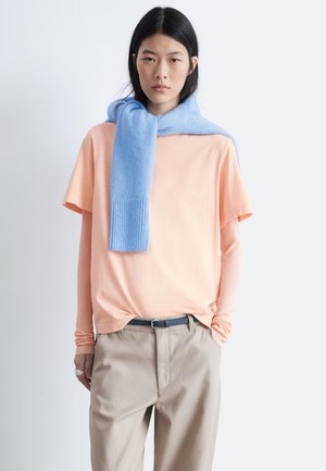 Pfirsichfarbenes Kurzarmoberteil über einem Langarmshirt, kombiniert mit beigen Hosen. Ein hellblauer Schal liegt locker um den Hals.