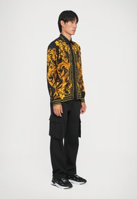Maglietta nera a maniche lunghe con un motivo dorato e testo, abbinata a pantaloni cargo neri e sneakers nere con accenti bianchi.