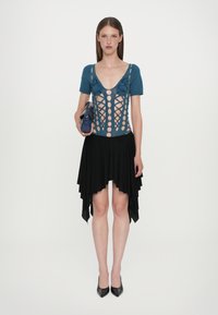 Τιρκουάζ ολόσωμο με κοψίματα και λεπτομέρειες με κορδόνια σε συνδυασμό με μια μαύρη ασύμμετρη φούστα. Το μοντέλο κρατά μια μπλε τσάντα. Μαύρες γόβες ολοκληρώνουν την εμφάνιση.