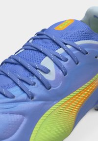 Κοντινό πλάνο μπλε αθλητικού παπουτσιού με υφασμάτινο επάνω μέρος με υφή, μπλε κορδόνια και κίτρινο-πορτοκαλί λεπτομέρεια στην ελαστική σόλα.