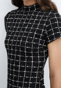 Mujer vistiendo una blusa de manga corta a cuadros en blanco y negro con un cuello alto, que muestra el hombro y la parte superior del torso.