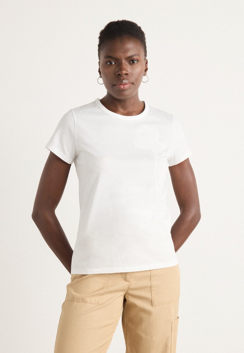 White cotton t-shirt with short sleeves, round neckline, and subtle floral design in the front. Worn with beige pants.