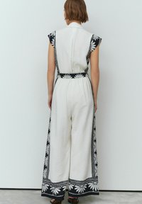 Vit jumpsuit med korta ärmar, prydd med svarta blommönster längs sidorna och nederkanten, åtdragen vid midjan för en avslappnad passform.
