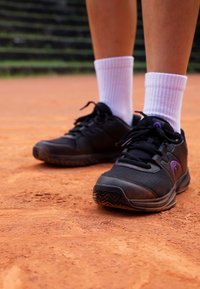 Black athletic shoes with purple accents, textured mesh upper, and rubber soles, paired with white ankle socks on a dirt surface.