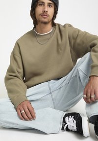 Un sweat-shirt olive à col rond et poignets côtelés, associé à un jean oversized bleu clair. Sneakers noires avec des lacets blancs et des anneaux en argent.