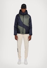 Quiltet jakke i marineblå og oliven med et diagonalt design, med hette og glidelås. Kombinert med kremfargede joggebukser og hvite sneakers.