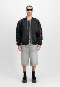 Black bomber jacket with a zip closure and patch detail, paired with gray denim shorts and black footwear. Model wears stylish sunglasses.