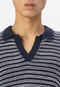 Polo en tricot rayé bleu marine et blanc avec un col en V et un col contrastant bleu marine. Texture douce avec des rayures horizontales sur toute la surface.