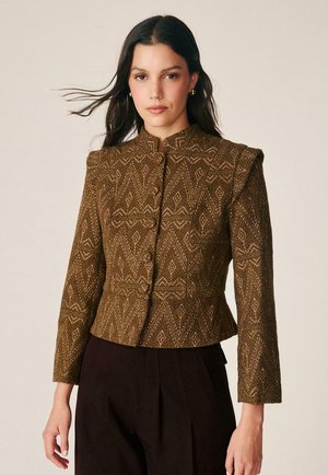 Mujer con chaqueta ajustada marrón con estampado y detalles en los hombros y pantalones negros sobre un fondo beige liso.
