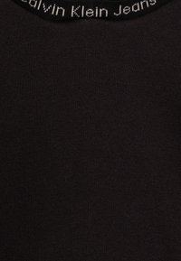 Tessuto di cotone nero con una vista ravvicinata del colletto, che presenta il marchio "Calvin Klein Jeans" in lettere bianche su un colletto a coste.