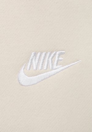 Beiger Stoff mit einer weichen Textur zeigt ein gesticktes weißes Nike-Logo, das von einem Swoosh-Design unter dem Markennamen hervorgehoben wird.