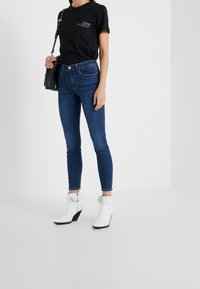 Camiseta de algodón negra con texto, combinada con jeans slim fit azules. El modelo lleva botas blancas hasta el tobillo con tacón cuña y detalle de hebilla plateada.