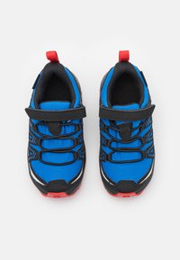 Zapatillas deportivas azules con acentos negros, parte superior texturizada, cordones elásticos y una correa de Velcro; lengüeta roja en el talón.