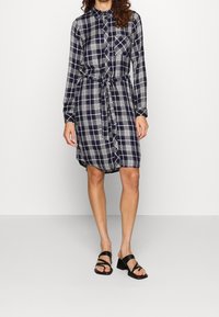 Donna che indossa un vestito a camicia a scacchi blu navy e bianco, lunghezza ginocchio, con una cintura in tessuto, abbinato a sandali con tacco a blocco aperti neri.