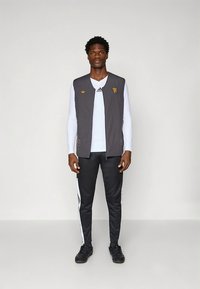 Γκρι αμάνικο γιλέκο με λογότυπο, φορεμένο πάνω από λευκό πουκάμισο με μακριά μανίκια. Μαύρη φόρμα με λευκές ρίγες και μαύρα αθλητικά παπούτσια.