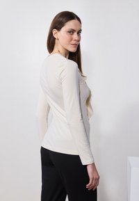 Long-sleeve cream top with a ruffled front detail, fitted shape, paired with black bottoms. Smooth texture and minimal design.