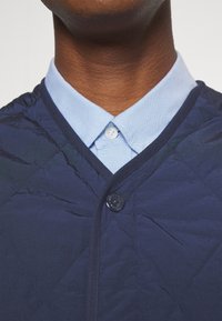 Navy gesteppte Weste mit V-Ausschnitt, ausgestattet mit einem blauen Kragen zum Knöpfen und einer abgerundeten Knopfdetail auf der Vorderseite.