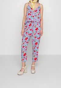 Combinaison florale en tissu bleu clair avec des fleurs rouges et roses ; pantalons courts ; bretelles réglables ; associée à des chaussures à talons carrés crème.